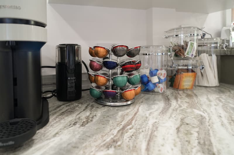A spinning rack of colorful coffee pods next to an electric coffee maker and jars of condiments on a marble countertop.
