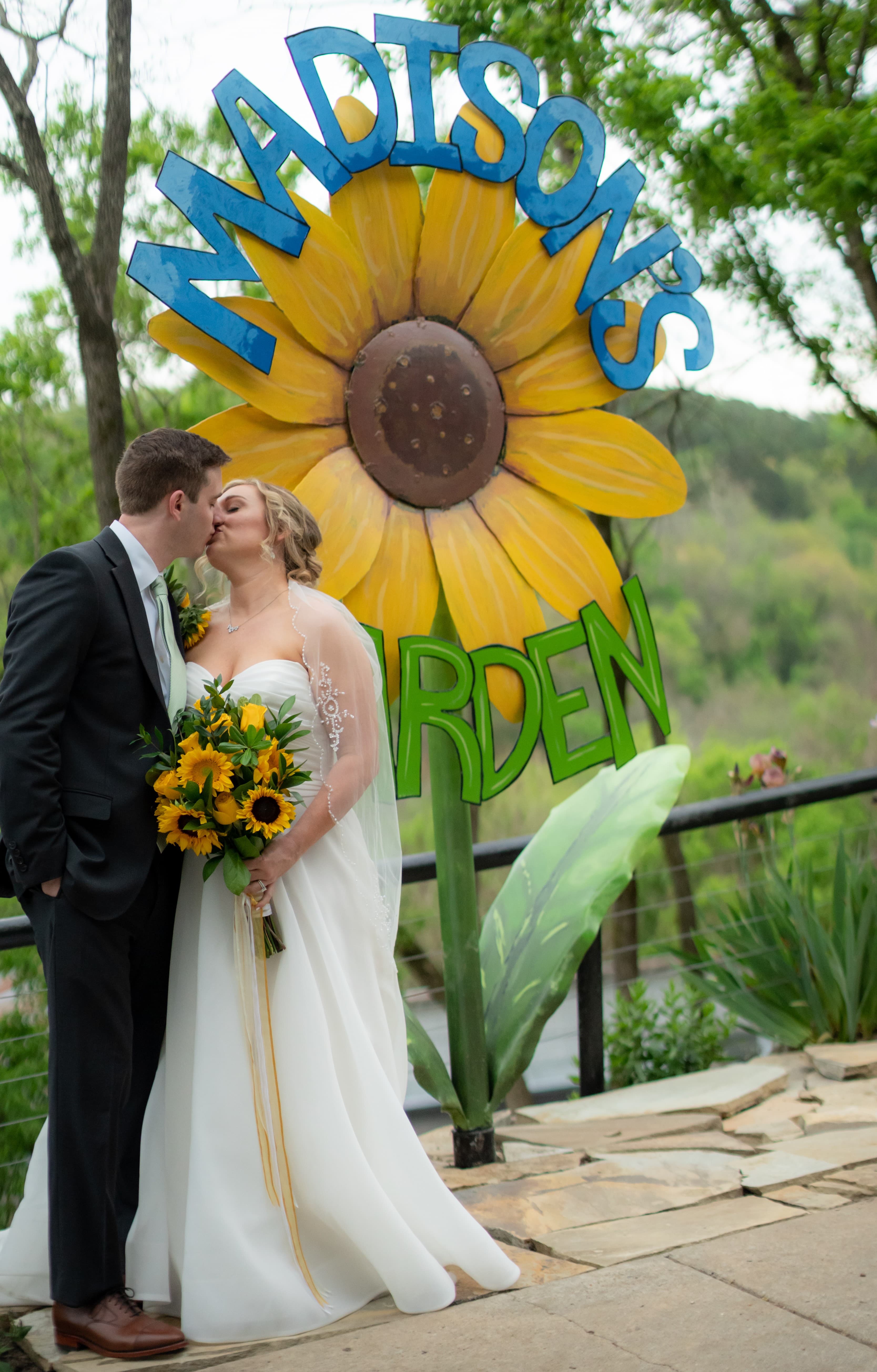 A newlywed couple kisses in front of a vibrant sunflower sign that reads "Madison's Garden."