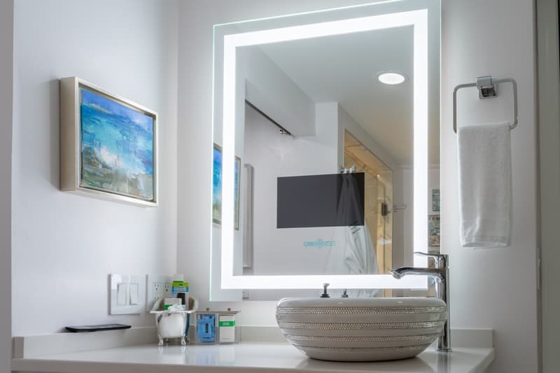 A modern bathroom sink area with a glowing mirror, a painting, and neatly arranged toiletries.