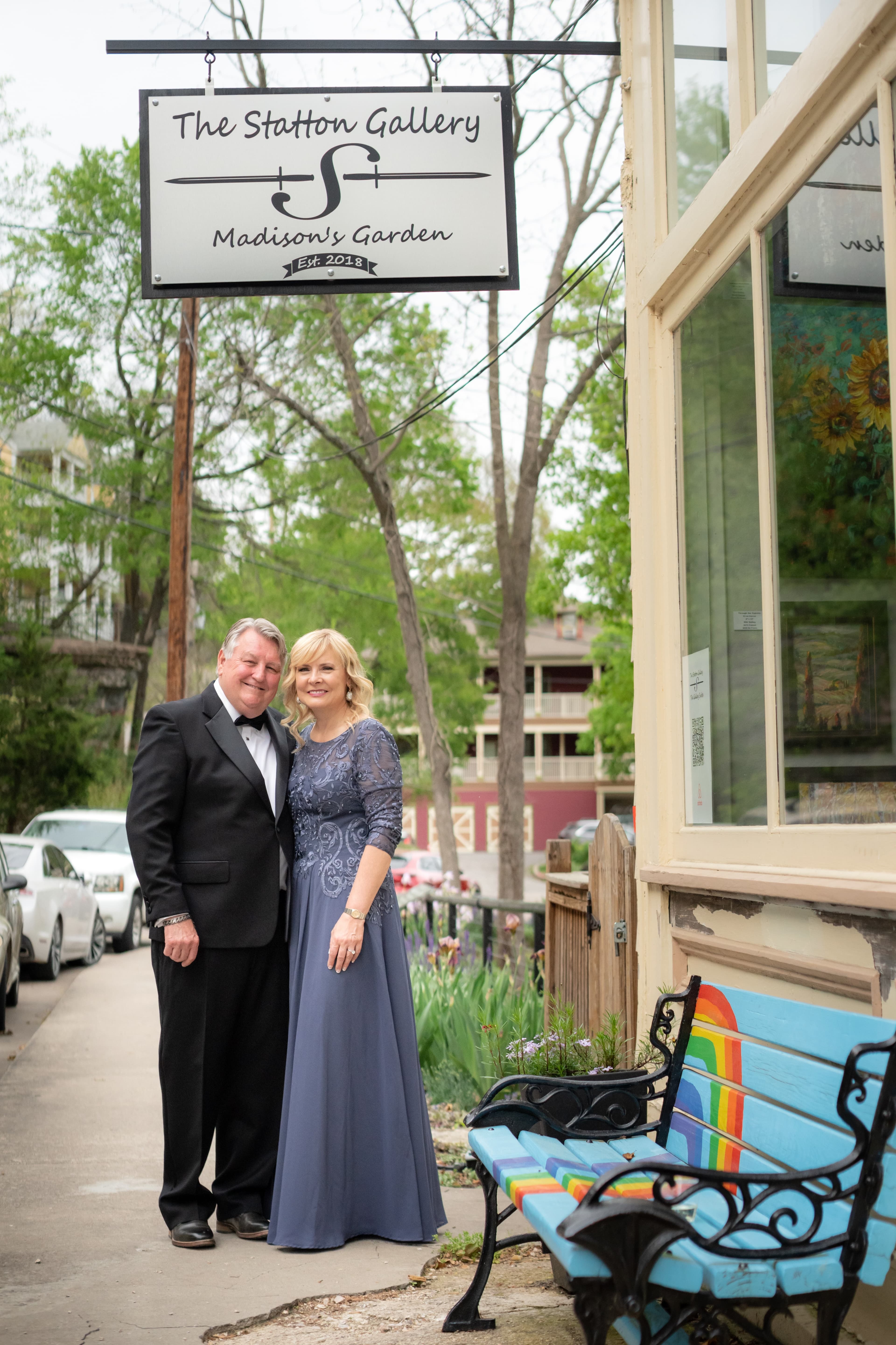 A couple in formal attire stands outside The Statton Gallery, smiling in front of a colorful bench and greenery.