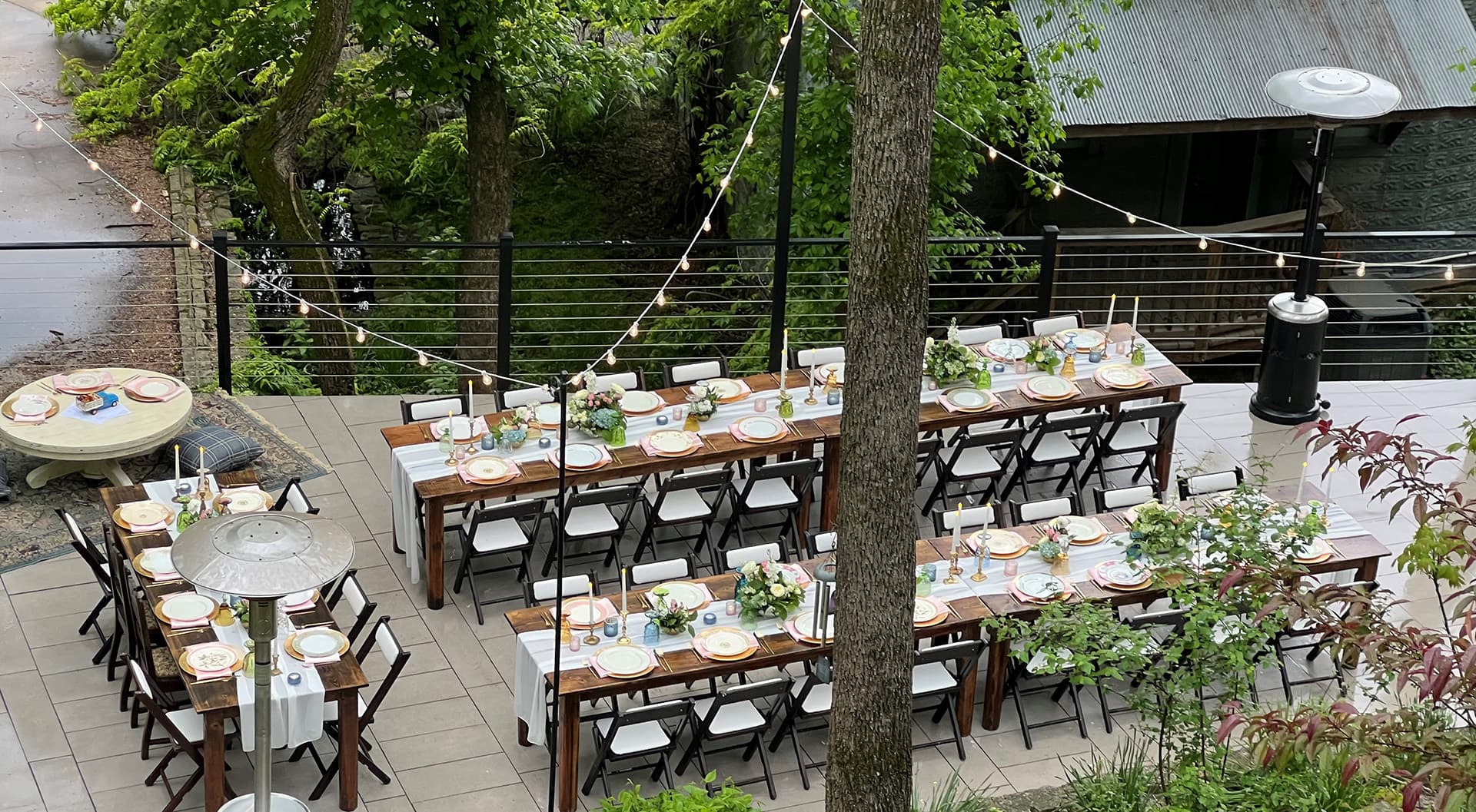 An outdoor dining setup featuring long wooden tables adorned with tableware and greenery under string lights.