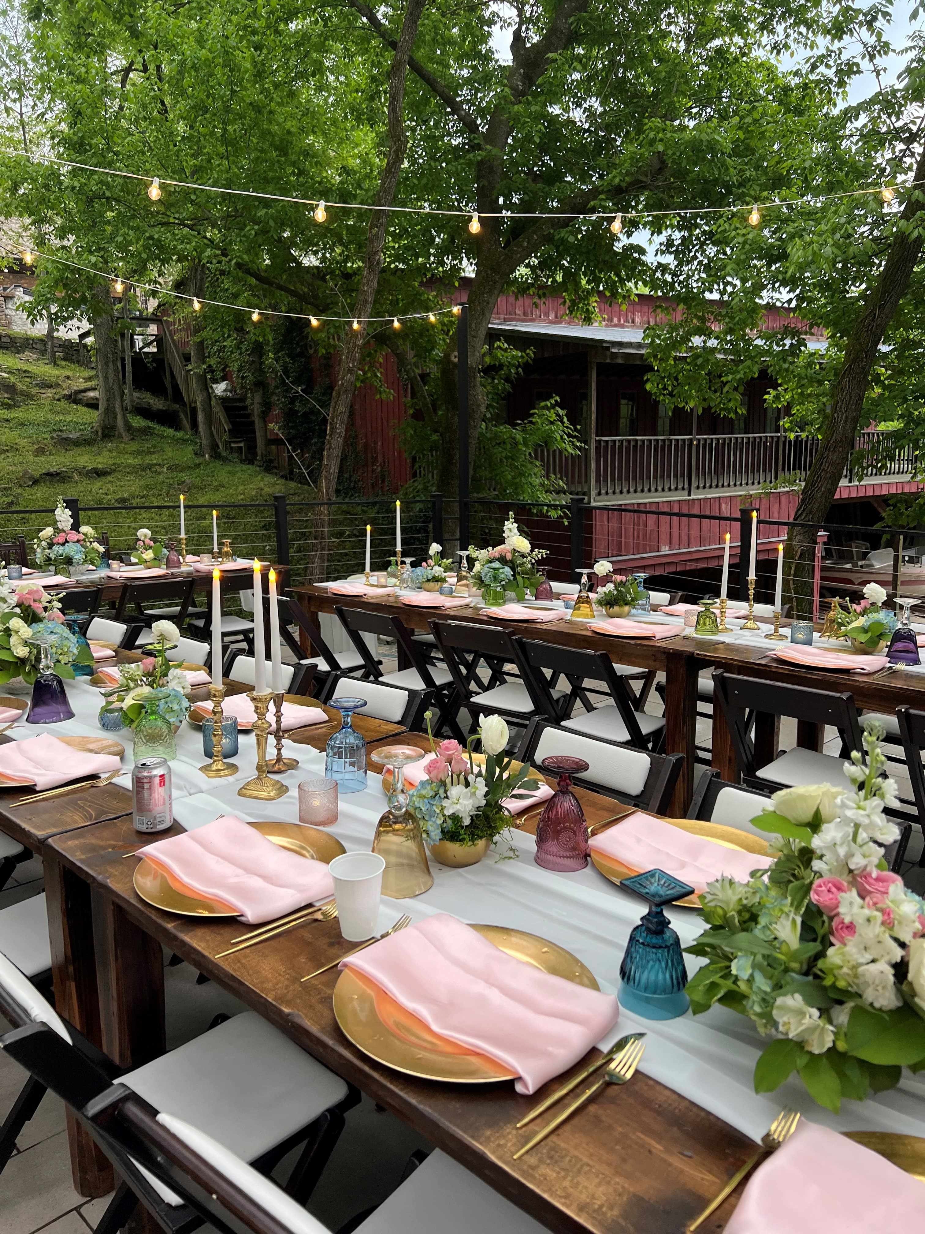 An elegantly set outdoor dining table with floral centerpieces, soft lighting, and pastel napkins.