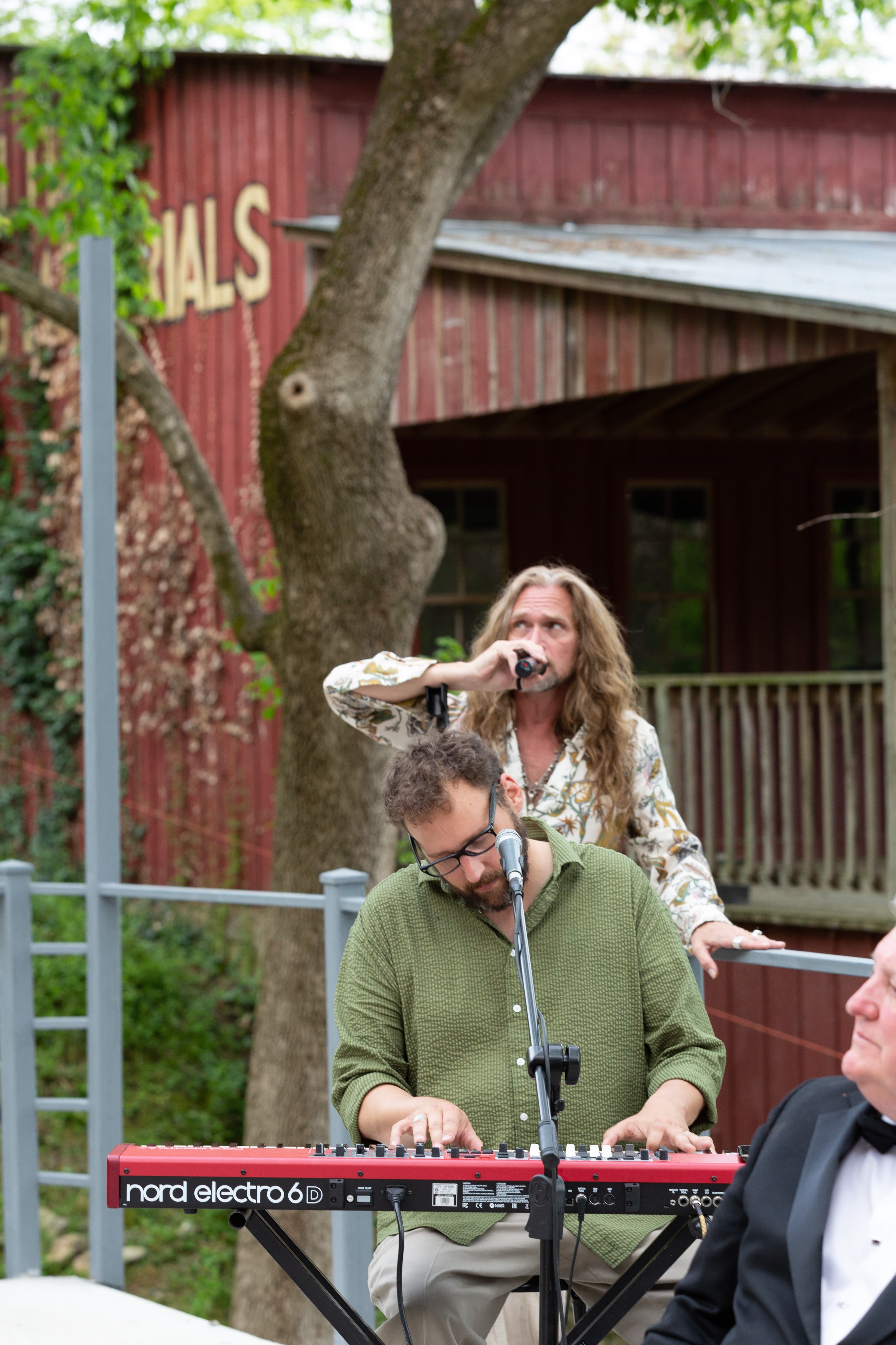 A musician plays a keyboard while another sings in a casual outdoor setting.