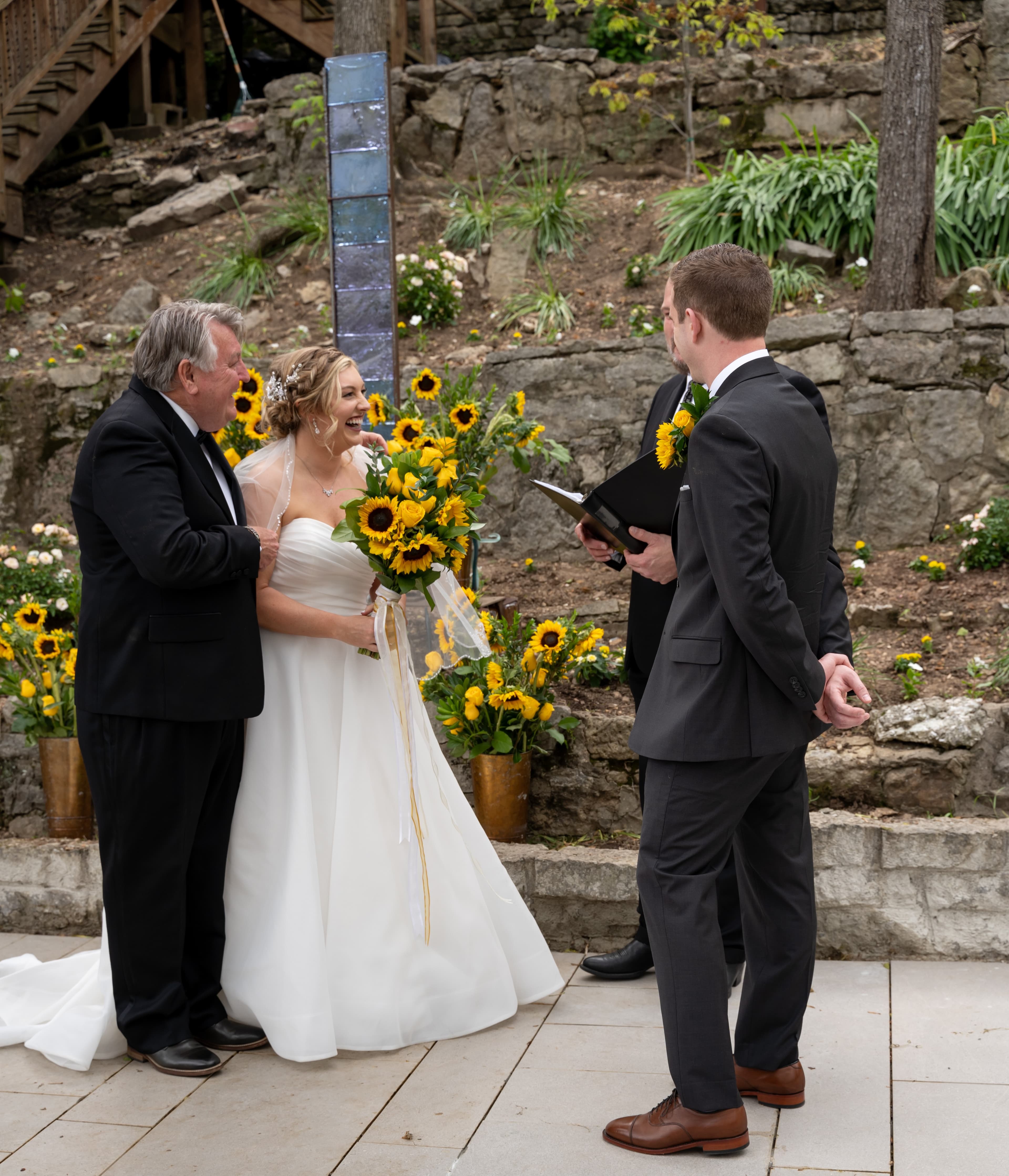 A bride holding sunflowers smiles at her officiant and father during their wedding ceremony outdoors.