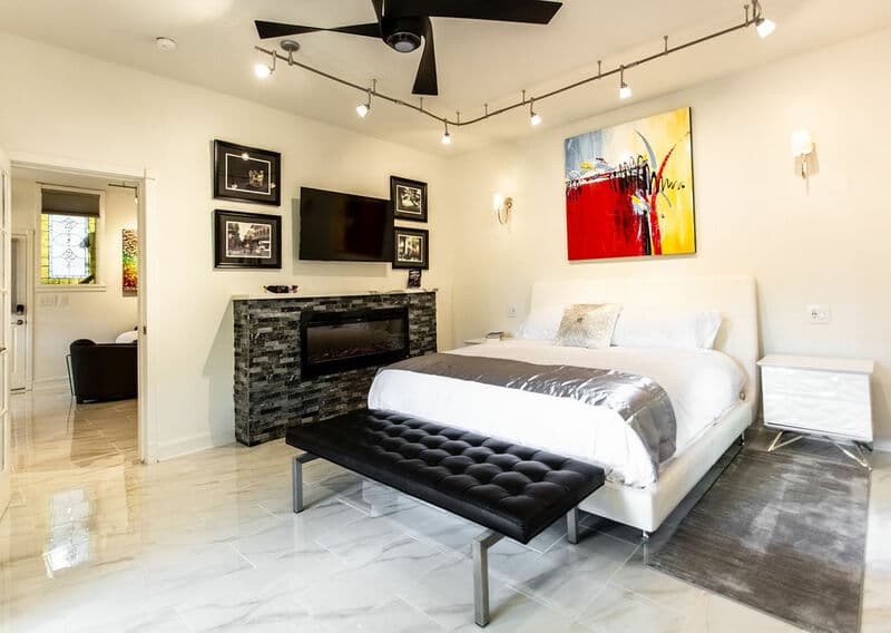 Modern bedroom featuring a large bed, a sleek bench, artwork, and a television on a stone accent wall.