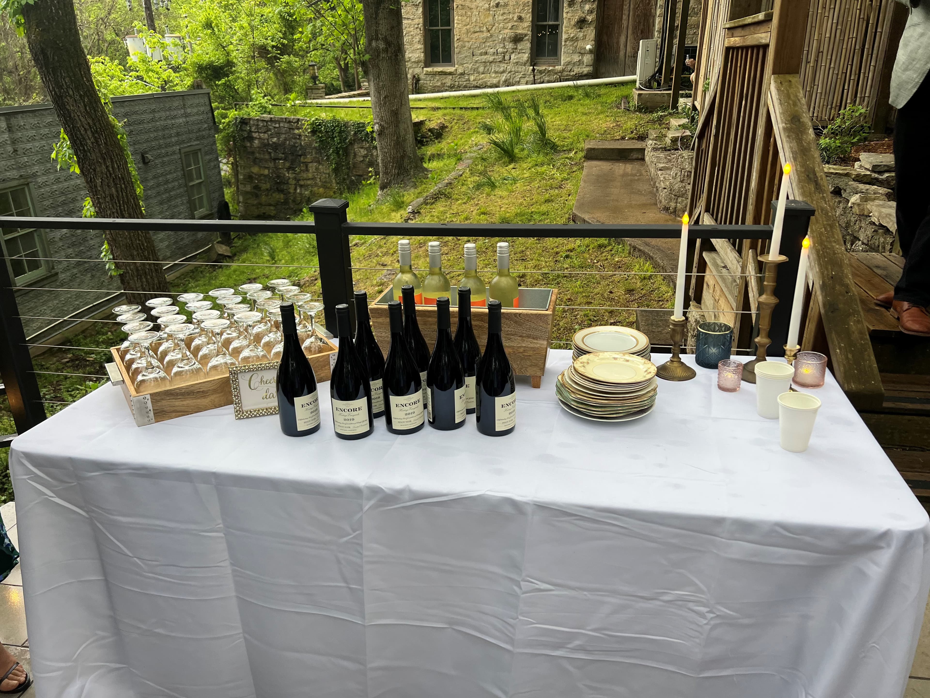 A white tablecloth displays wine bottles, glasses, plates, and candles outdoors.