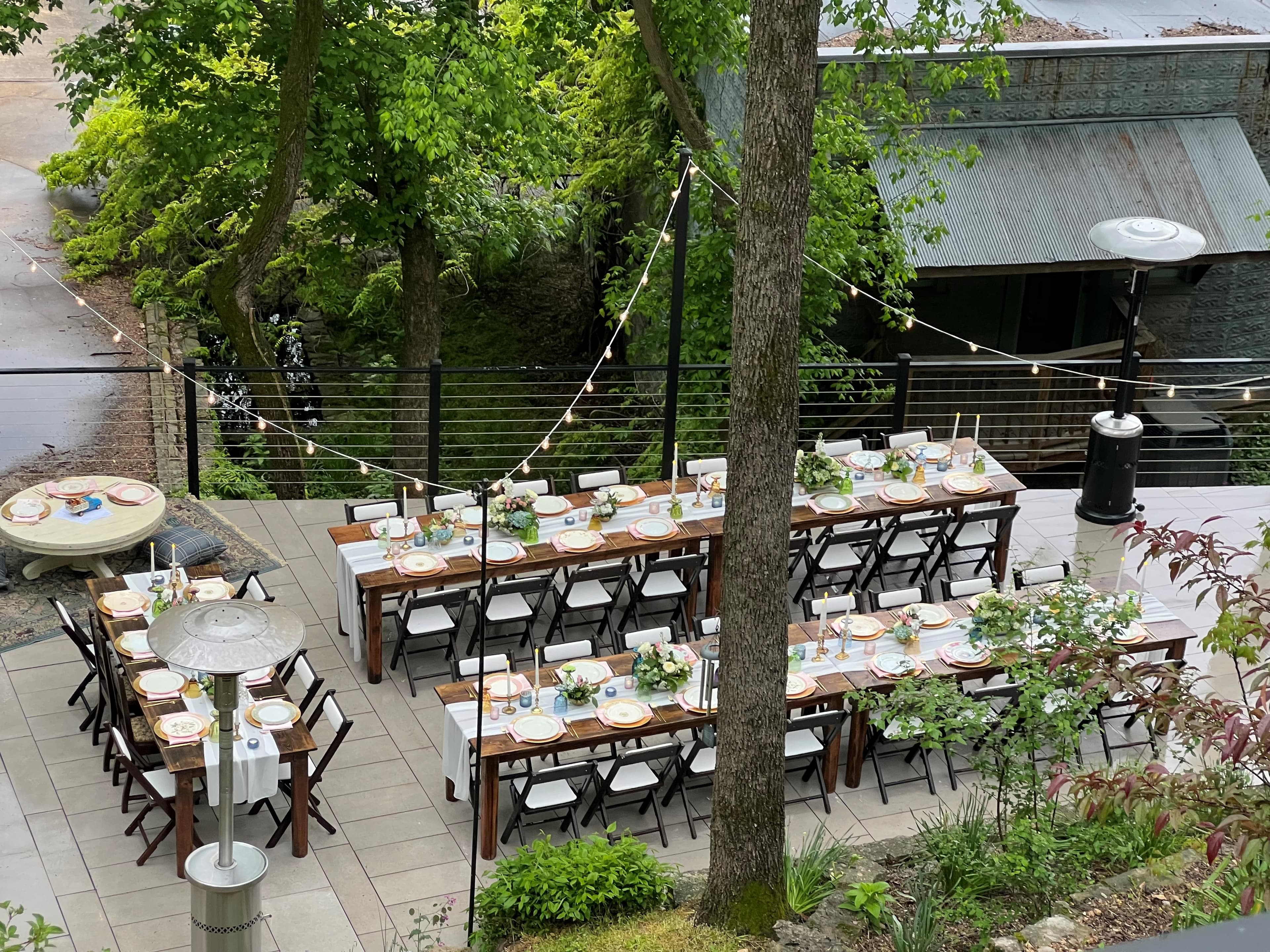 Outdoor dining setup with long tables, chairs, and decorative greenery under string lights.