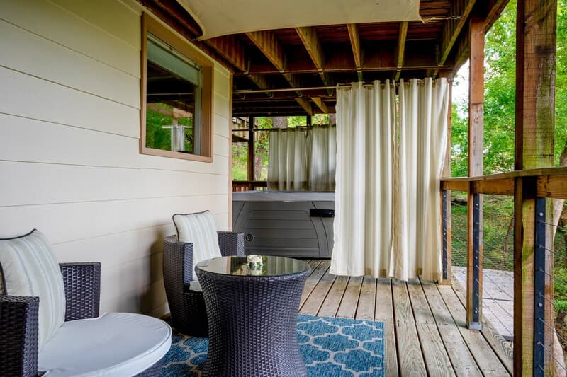 A cozy porch area featuring two chairs, a small round table, and curtains, surrounded by greenery.