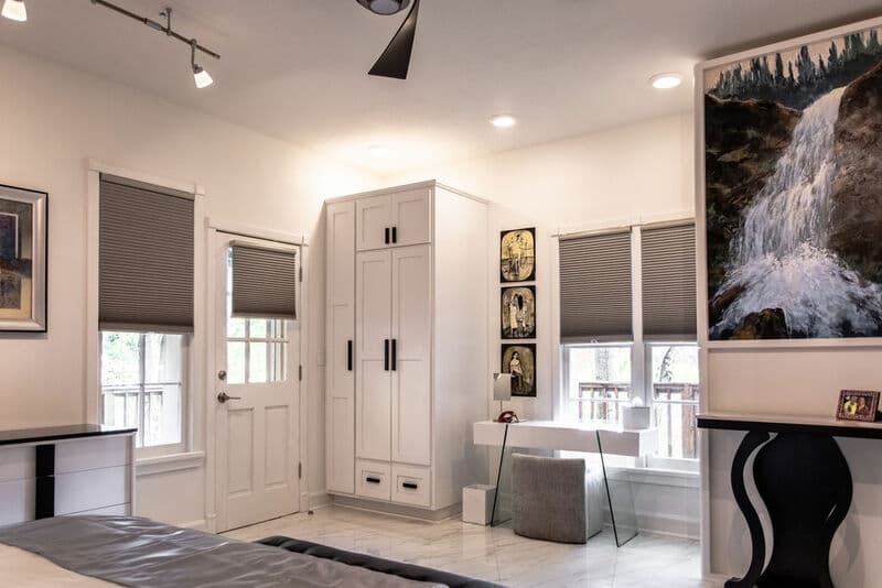 A modern bedroom featuring a white wardrobe, a glass desk, and artwork, with natural light coming through the windows.