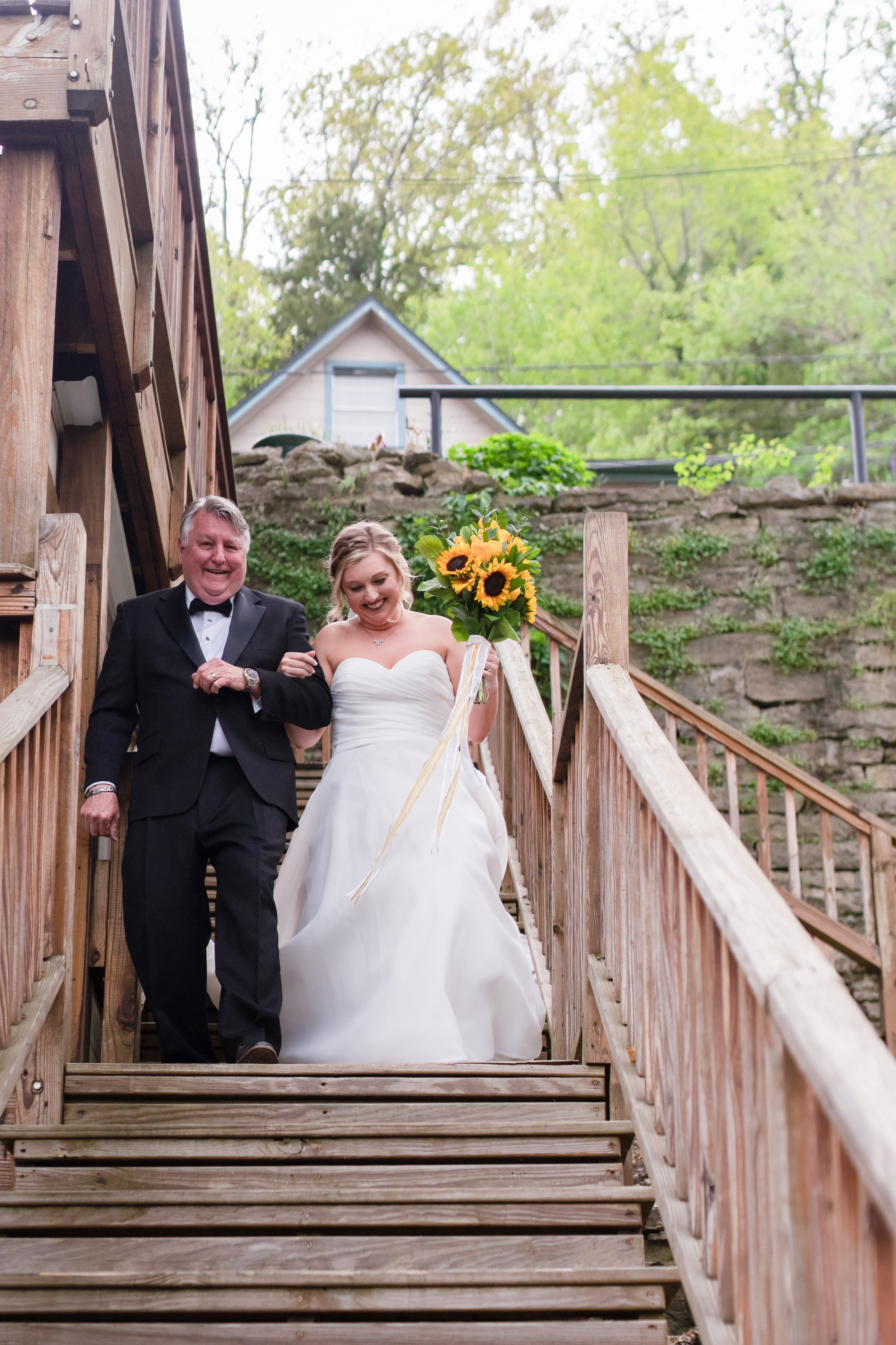A bride in a white gown smiles as she walks down wooden stairs with her father, who is wearing a tuxedo, holding a bouquet of sunflowers.